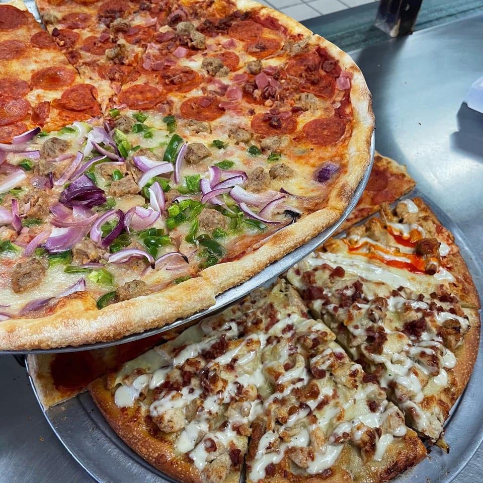 Two pizzas on metal trays, one with pepperoni, sausage, and ham, the other with chicken, bacon, and ranch.
